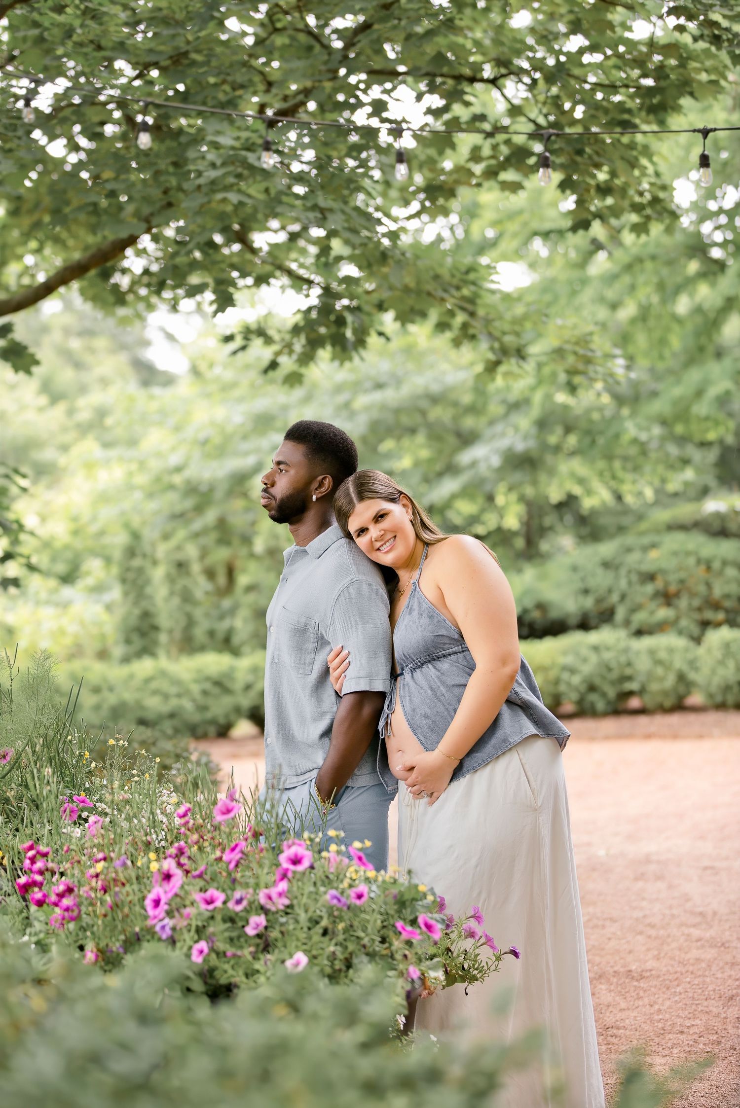 Couple in matching gray outfits posing in a lush garden with pink flowers and string lights overhead.