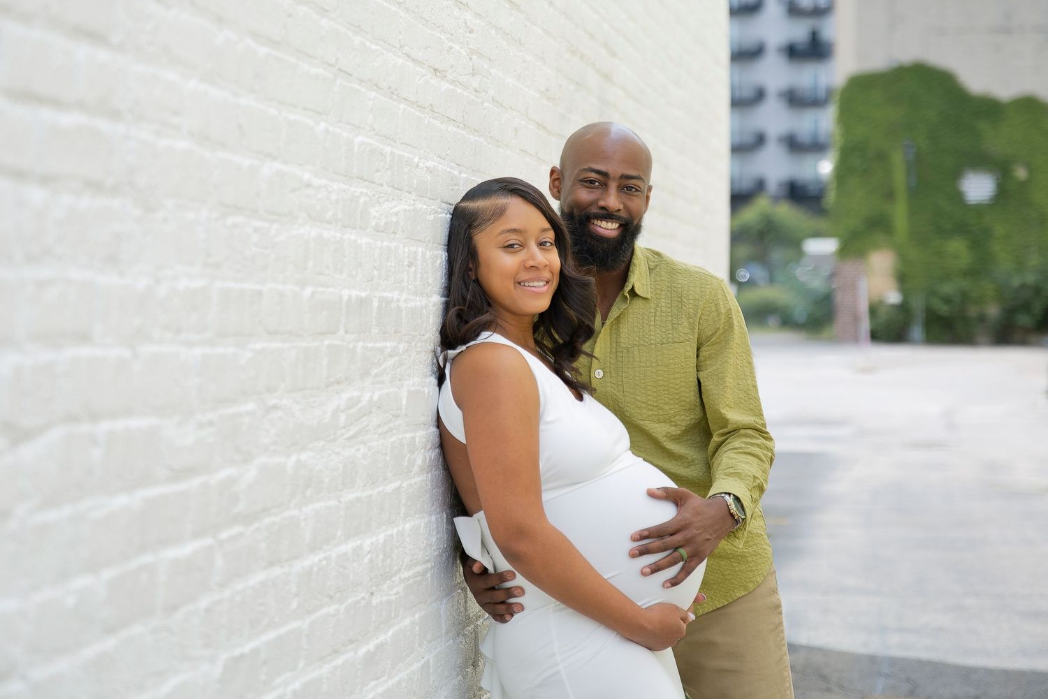 A smiling couple poses against a white brick wall, with the man tenderly holding the pregnant woman's belly.