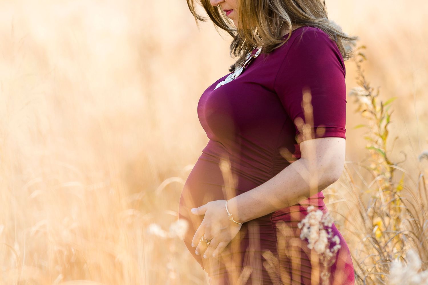 Woman in burgundy dress poses in golden wheat field during sunset with soft glowing light.