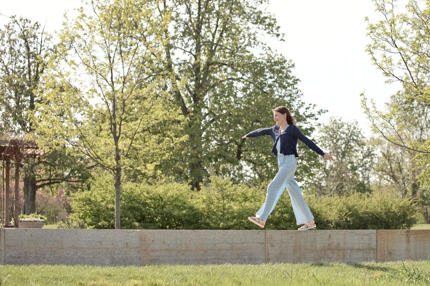 Young woman balancing on a low stone wall in a sunny park surrounded by green trees.
