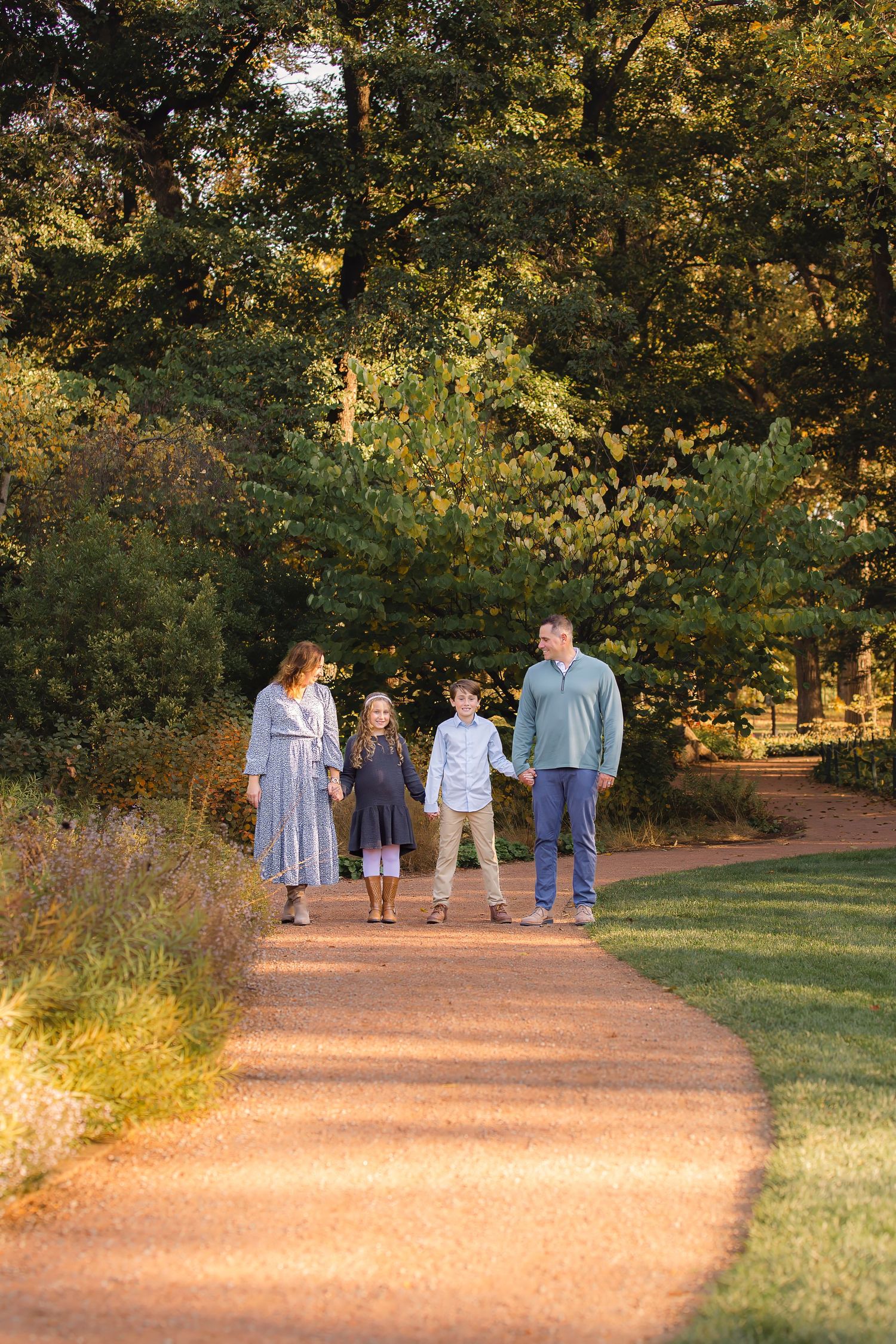 A group of people walking along a sunlit dirt path bordered by lush trees and grass in a park at golden hour.