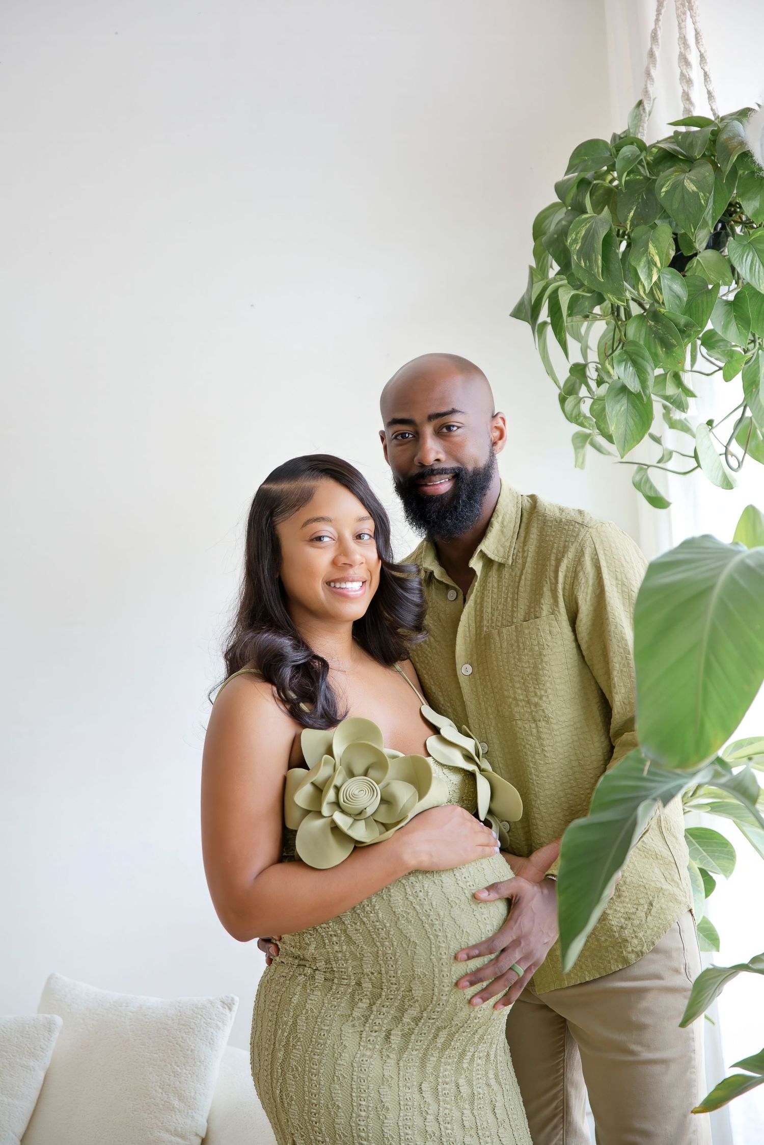Pregnant couple in matching green outfits posing together surrounded by lush houseplants in a bright studio.