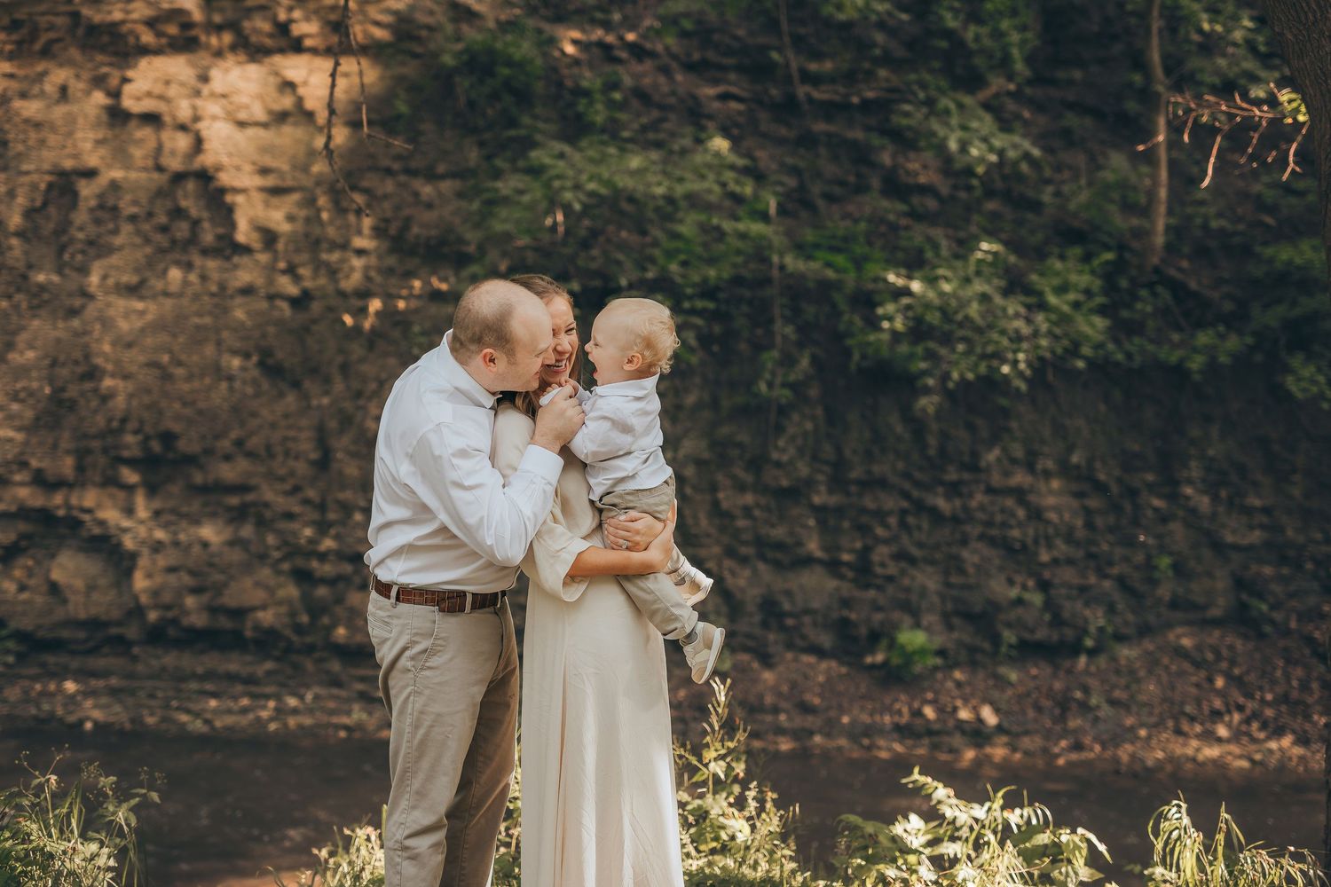 Couple sharing a tender moment with their baby outdoors near a rocky cliff and lush greenery.