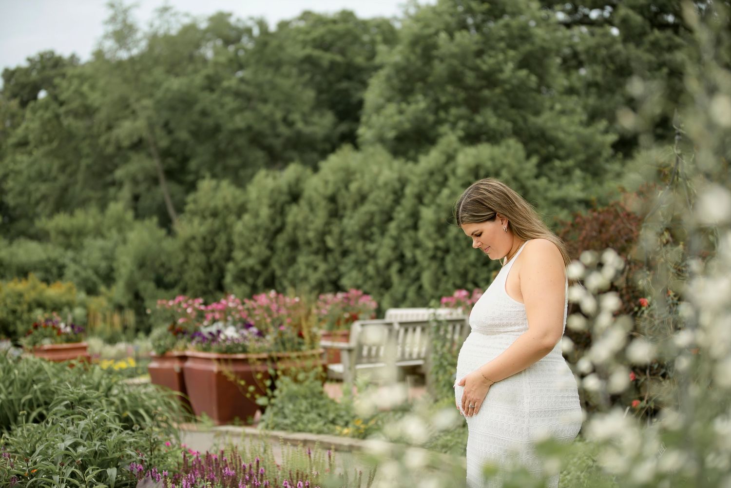 Pregnant woman in white dress standing in a lush garden, looking down at her baby bump.