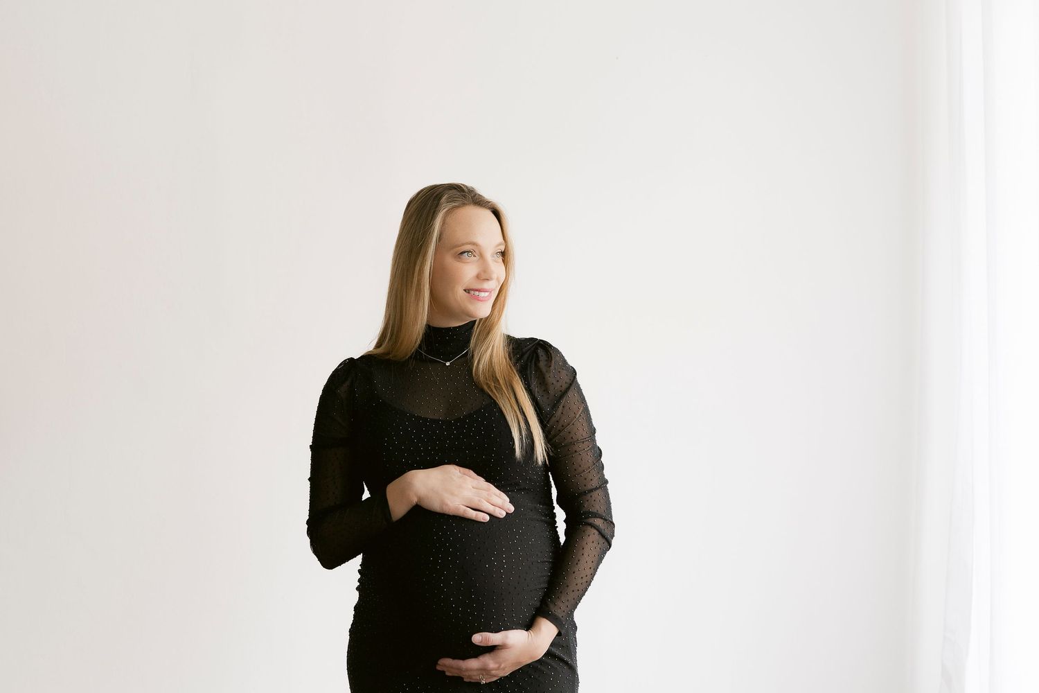 Pregnant woman in black dress cradling baby bump, smiling sideways in bright white studio maternity photo.