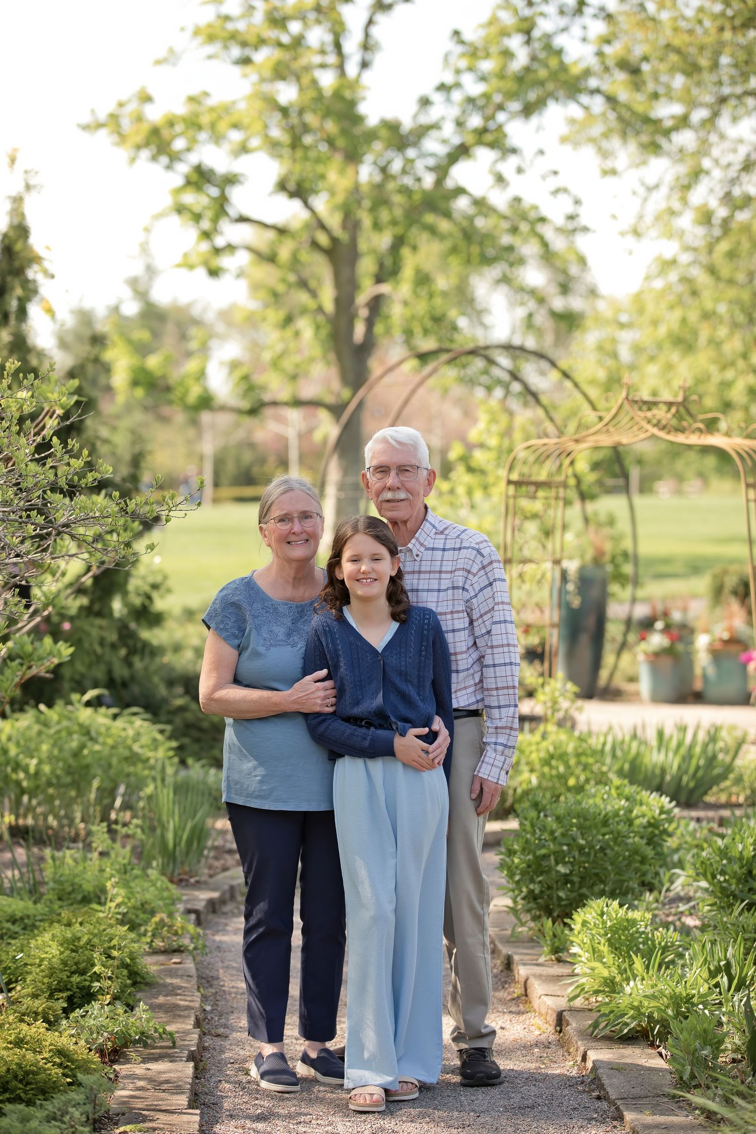 Grandparents and granddaughter posing on a garden path surrounded by greenery in warm golden hour light.