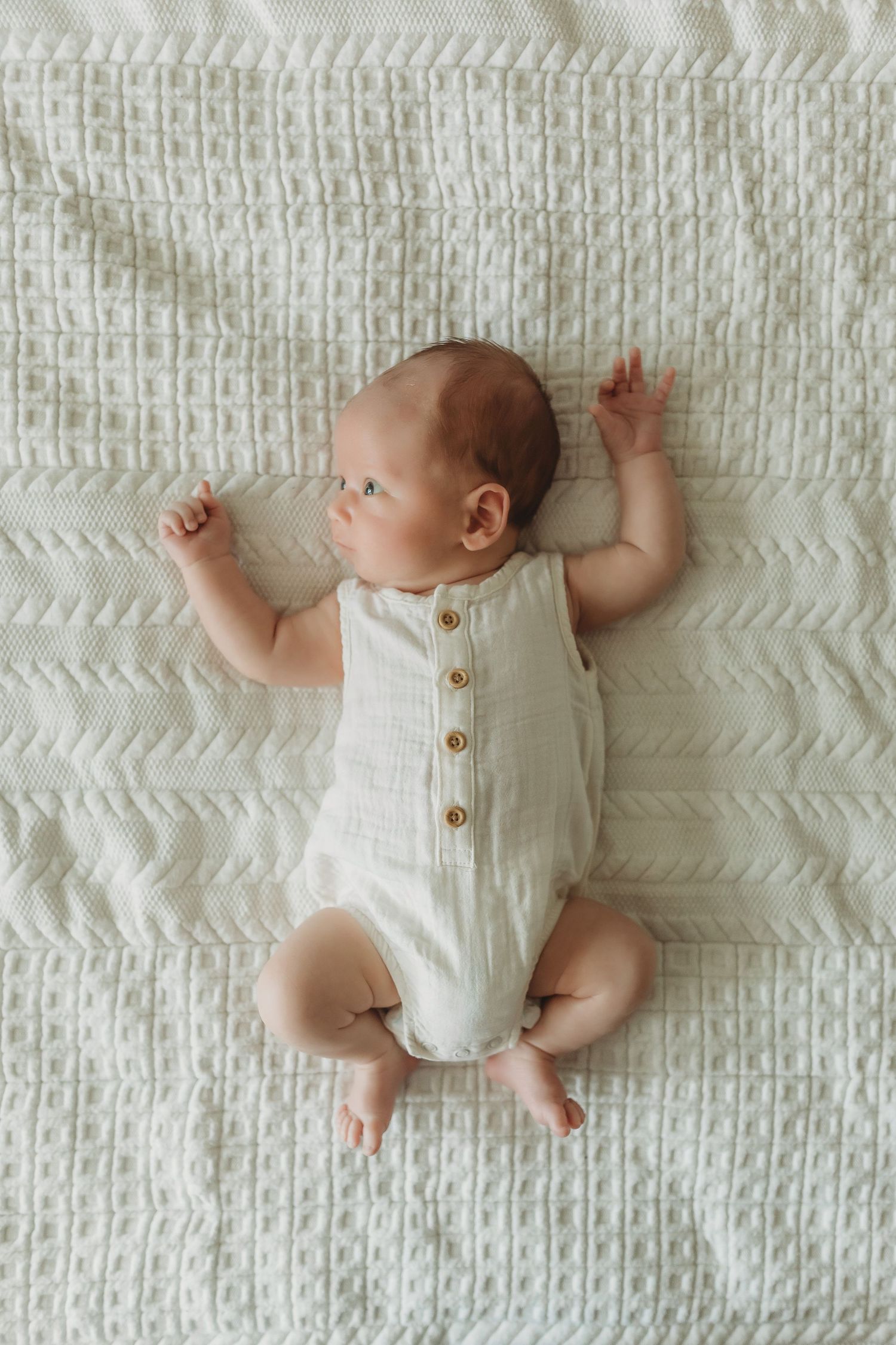 Overhead view of infant wearing white romper lying on textured white blanket.