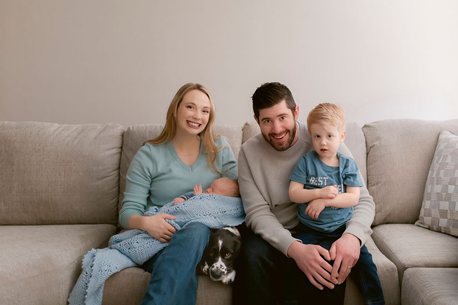 A family sits together on a gray couch with their small dog, sharing a relaxed moment at home.