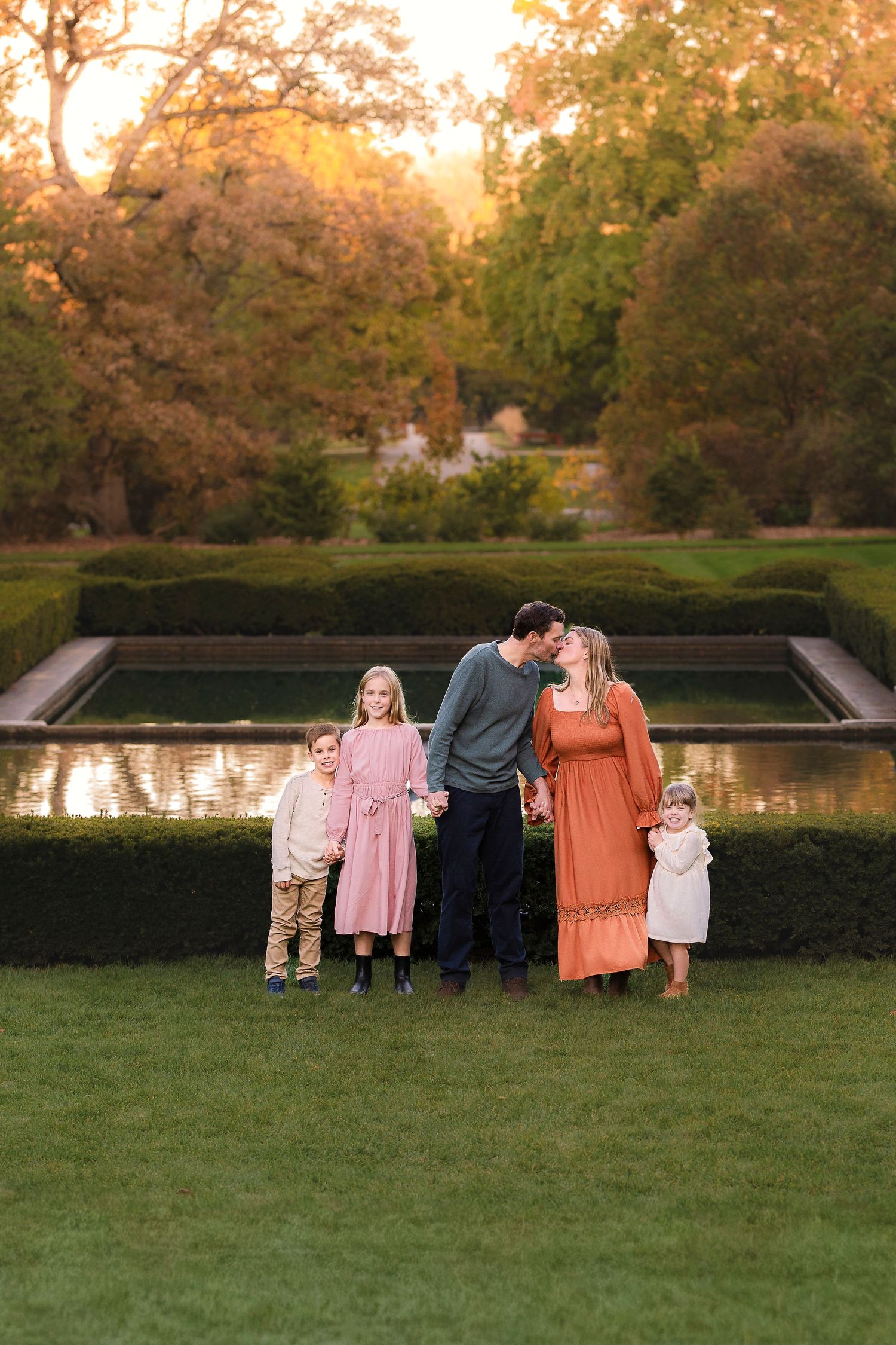 Family in coordinating outfits stands by a reflective pond at sunset in a manicured garden setting.