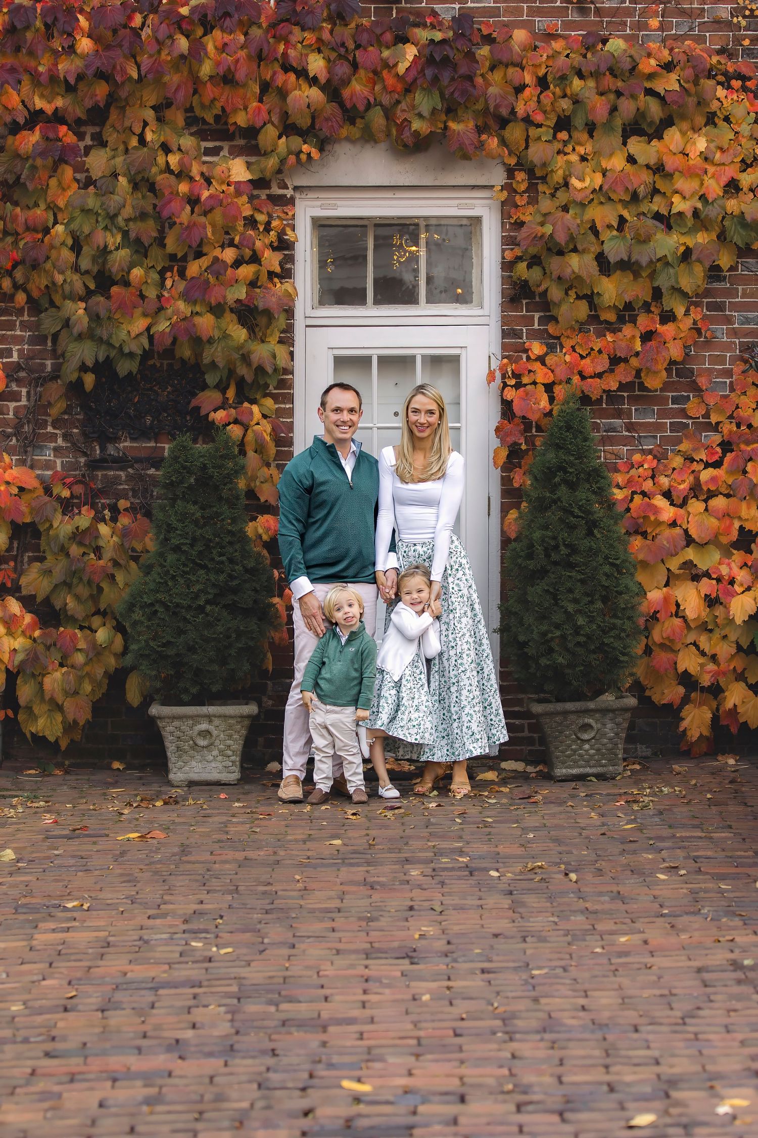 Family of four posing in front of brick wall covered in colorful autumn foliage beside a white door.