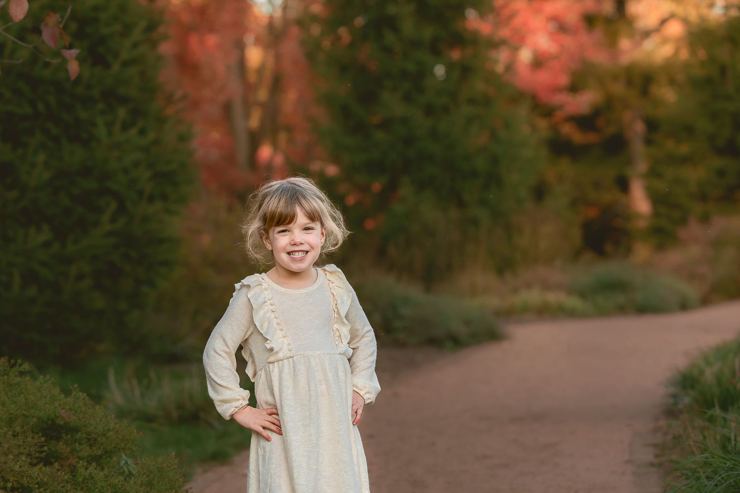 A child in a cream-colored dress walks along a wooded path during autumn with warm orange foliage in the background.