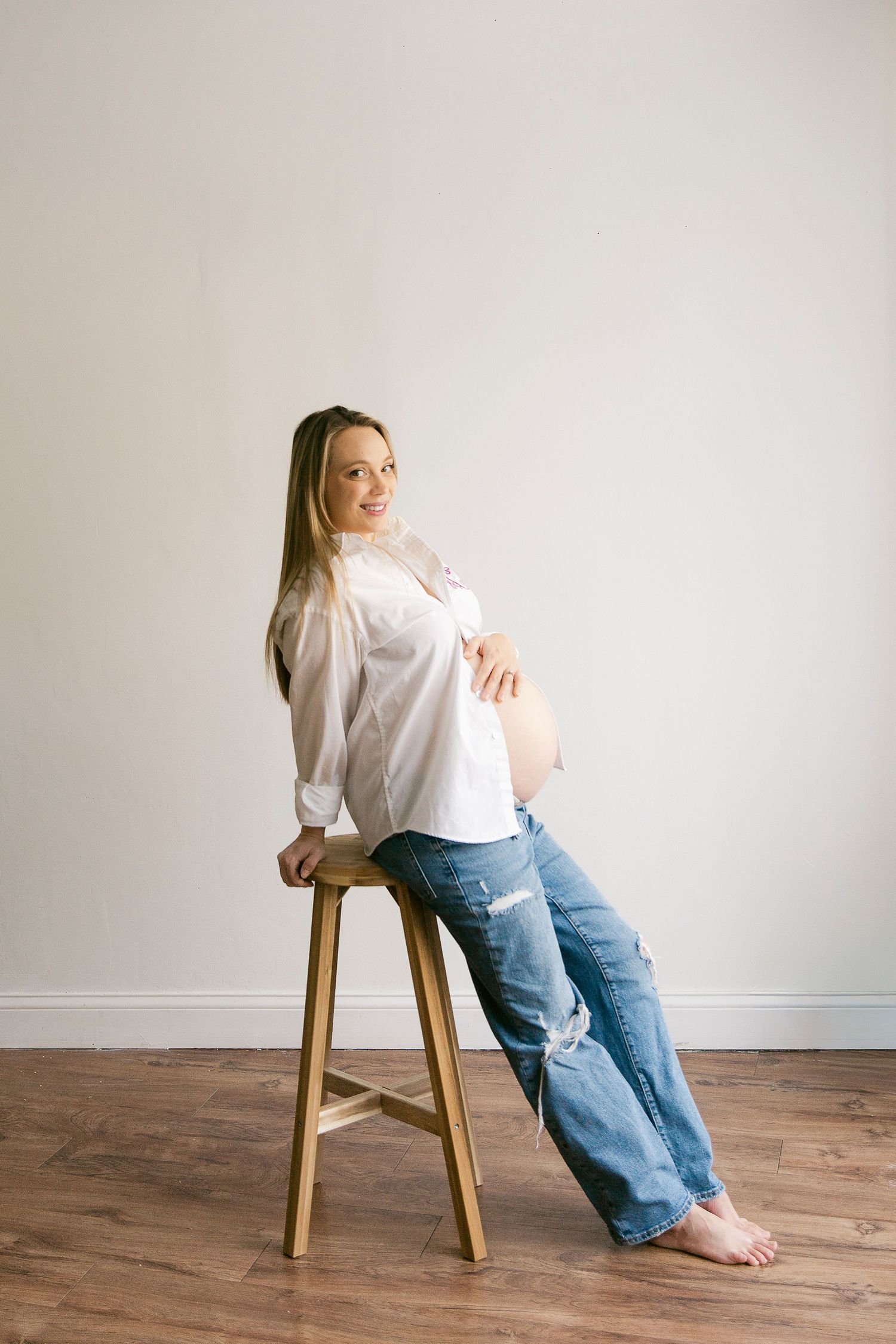 A person in a white shirt and blue jeans poses casually on a wooden stool against a minimalist white backdrop.