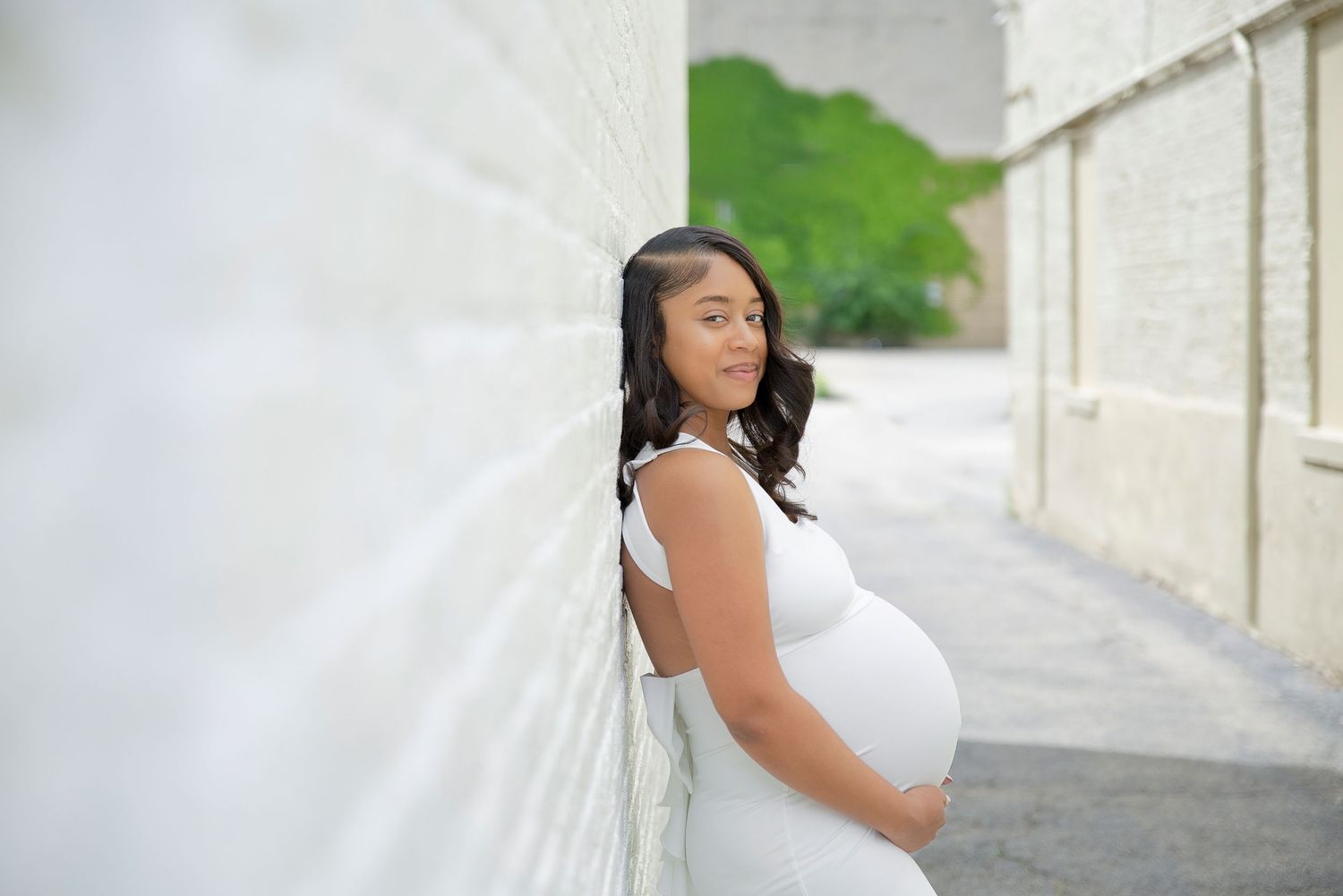 Pregnant woman in white dress leaning against white brick wall outdoors during maternity photo session.