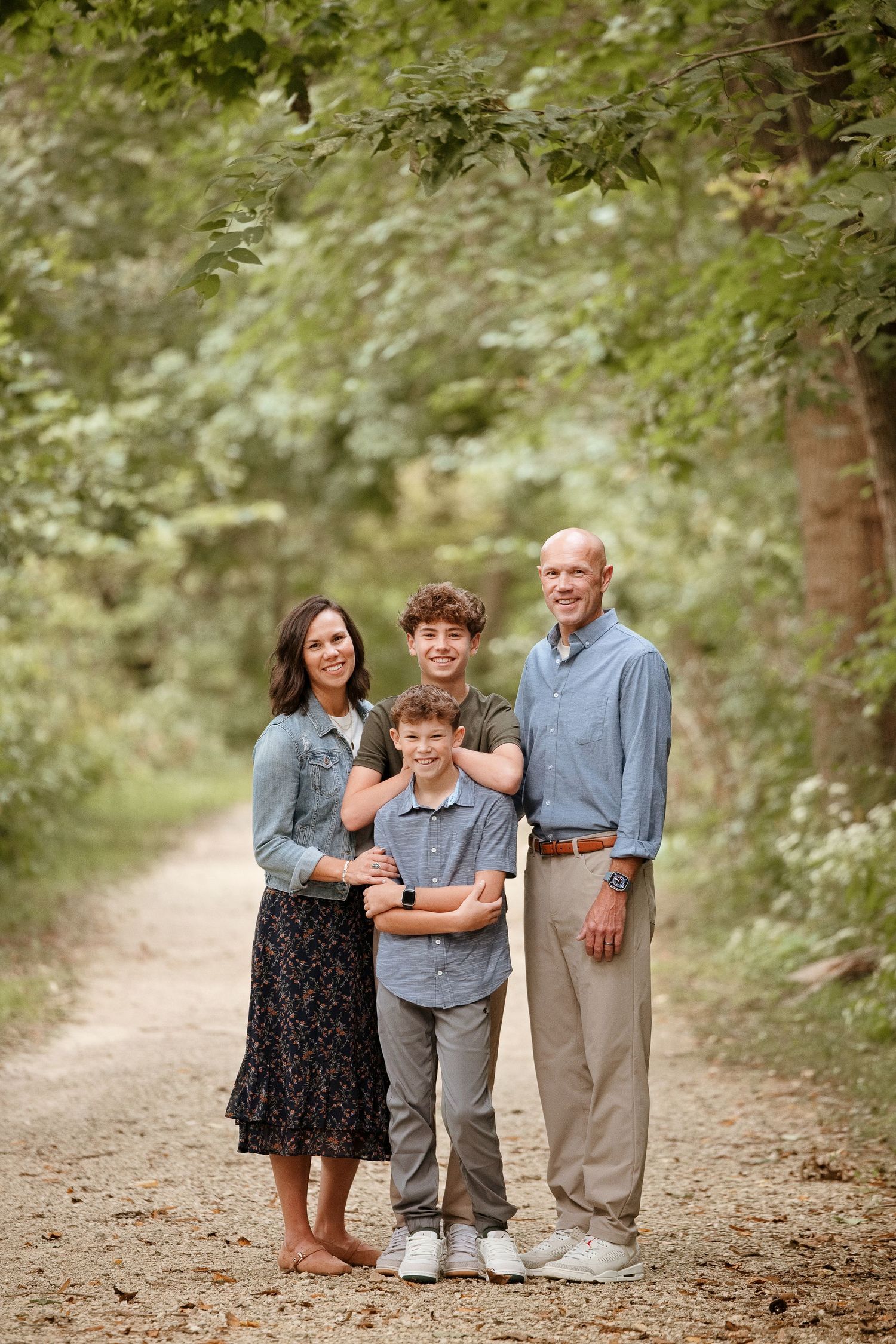 Family of four posing on a tree-lined path, parents with two boys smiling outdoors in casual attire.