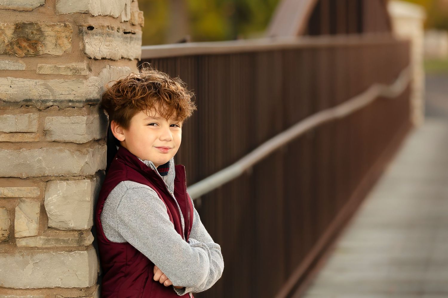 A child in a burgundy vest and gray sweater leans against a stone pillar on a sunny day.