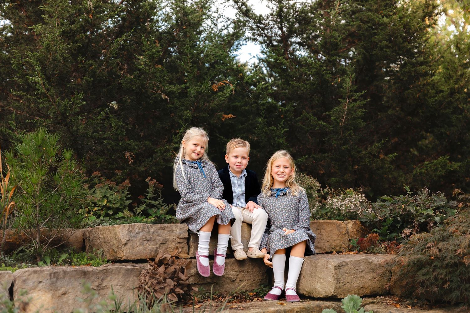 A group sits together on a stone wall surrounded by lush greenery in an outdoor garden setting.
