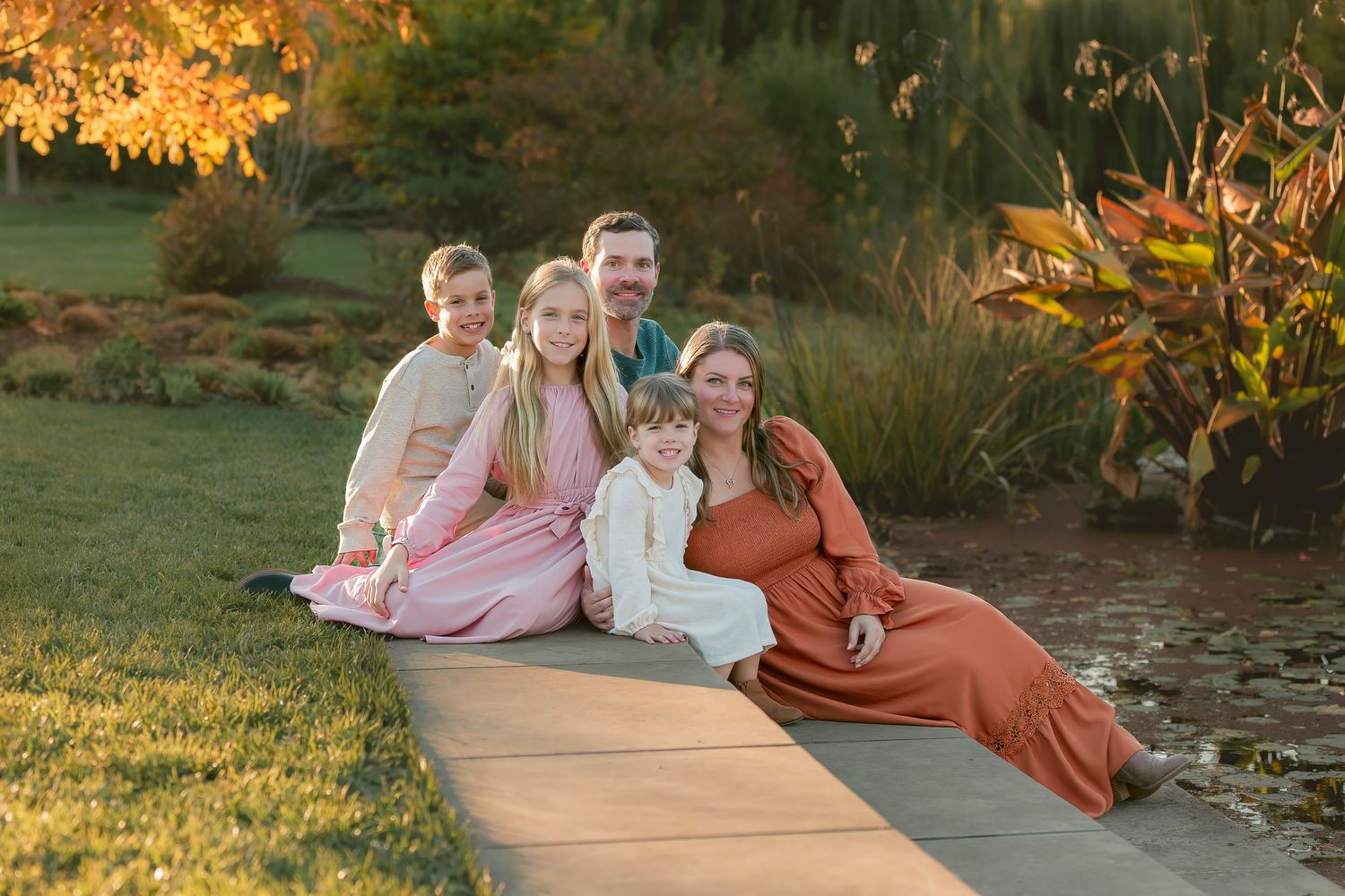 Family wearing coordinated fall outfits poses together on wooden bench during golden hour sunset photoshoot.
