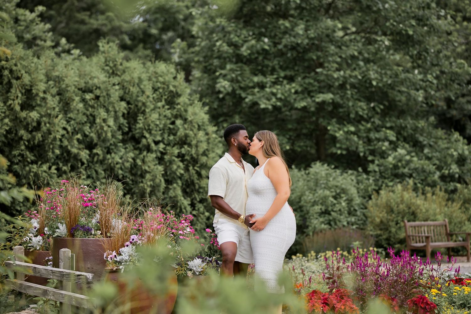 Interracial couple sharing a kiss in a lush garden, woman in white maternity dress, surrounded by colorful flowers.