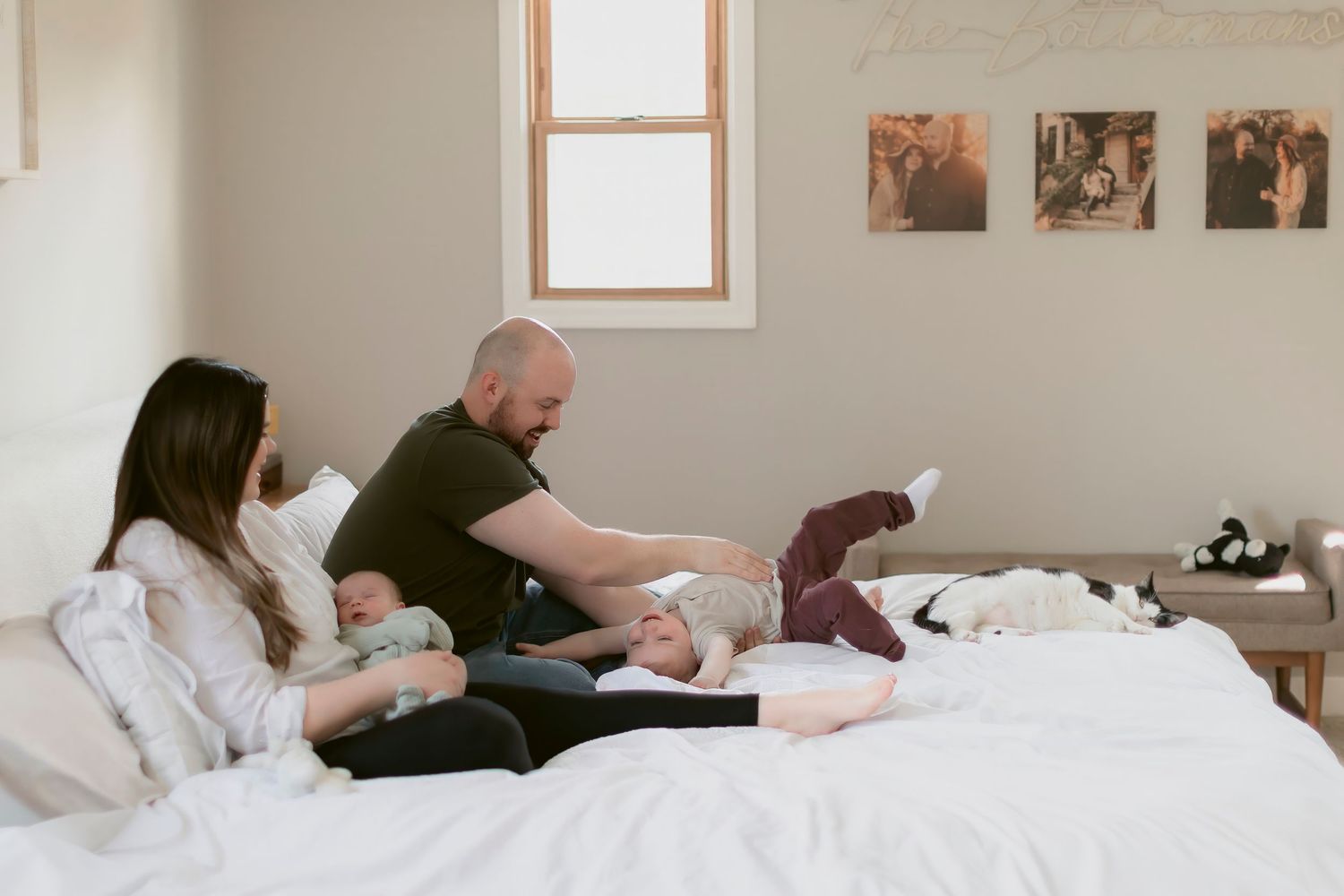 Family relaxing together on a white bed, with a bald man tending to a baby while a woman holds another child.