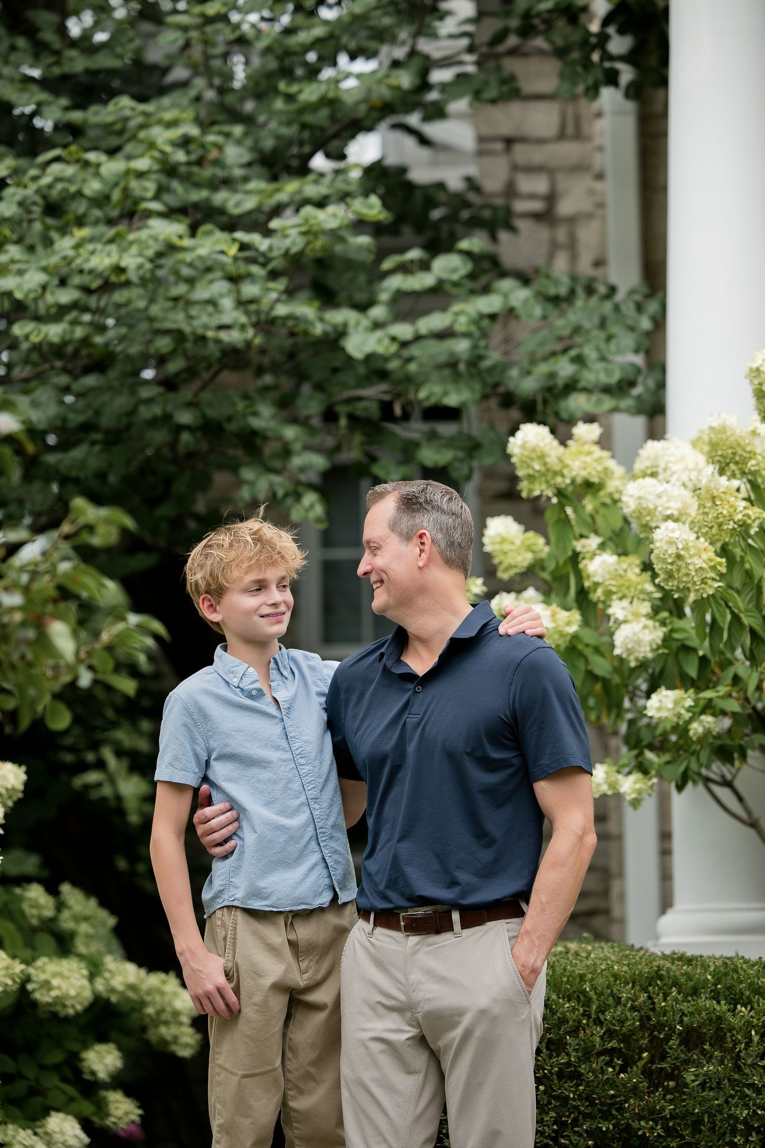 A smiling father and teenage son stand together outdoors near blooming hydrangeas and lush greenery.