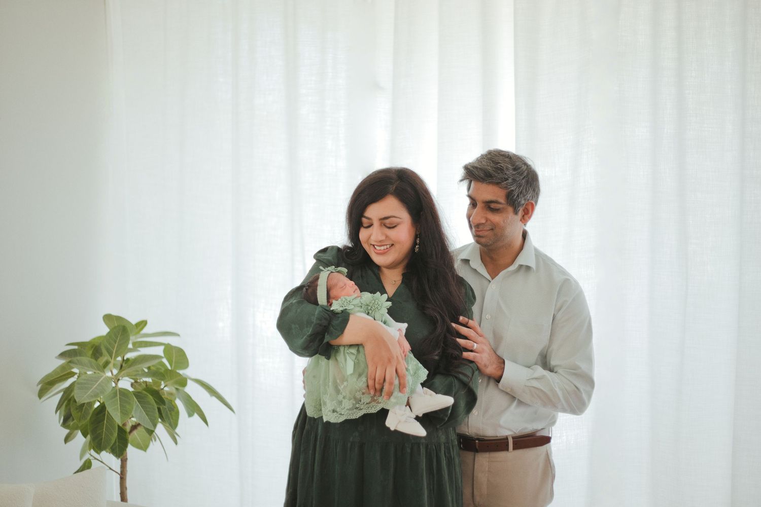 A couple smiles while cradling their newborn baby in a bright, airy room with white curtains and a green plant.
