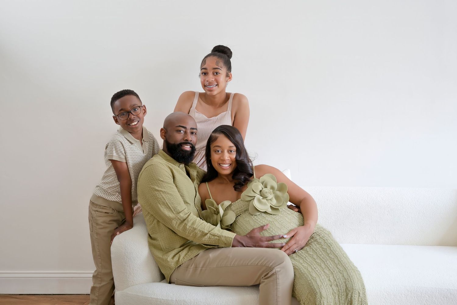 Pregnant Black woman poses with her family on a white sofa during a maternity photo session.