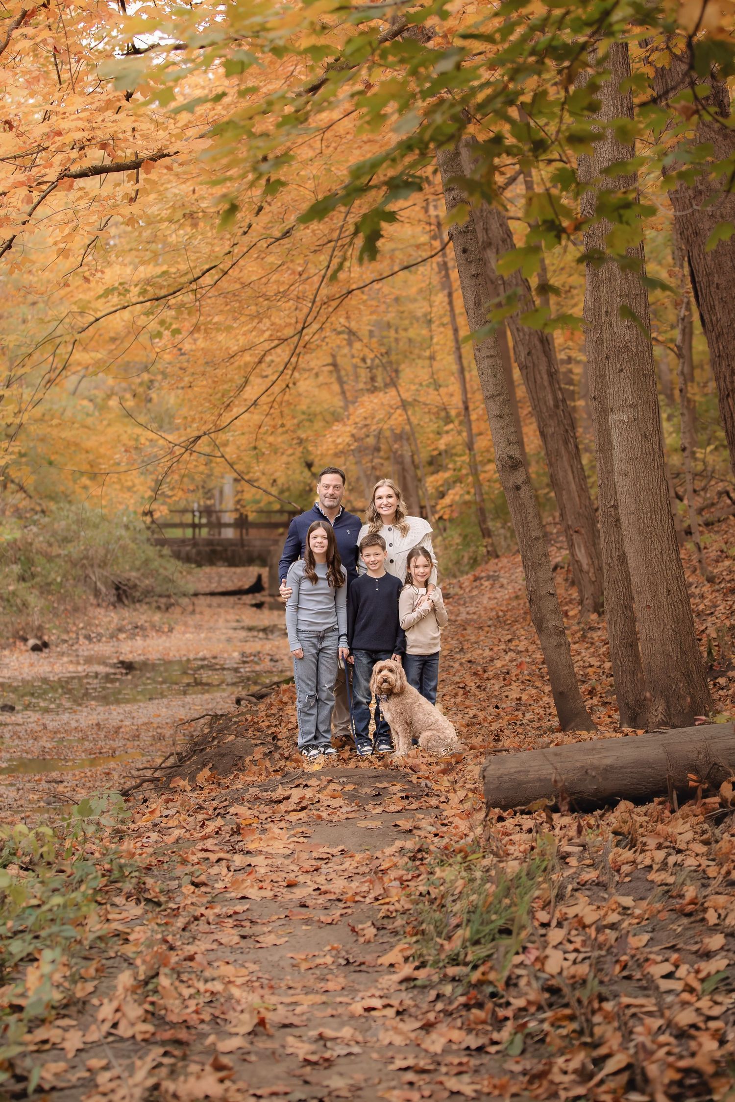 Family of five with a golden doodle dog posing on a leaf-covered forest path surrounded by vibrant autumn foliage.