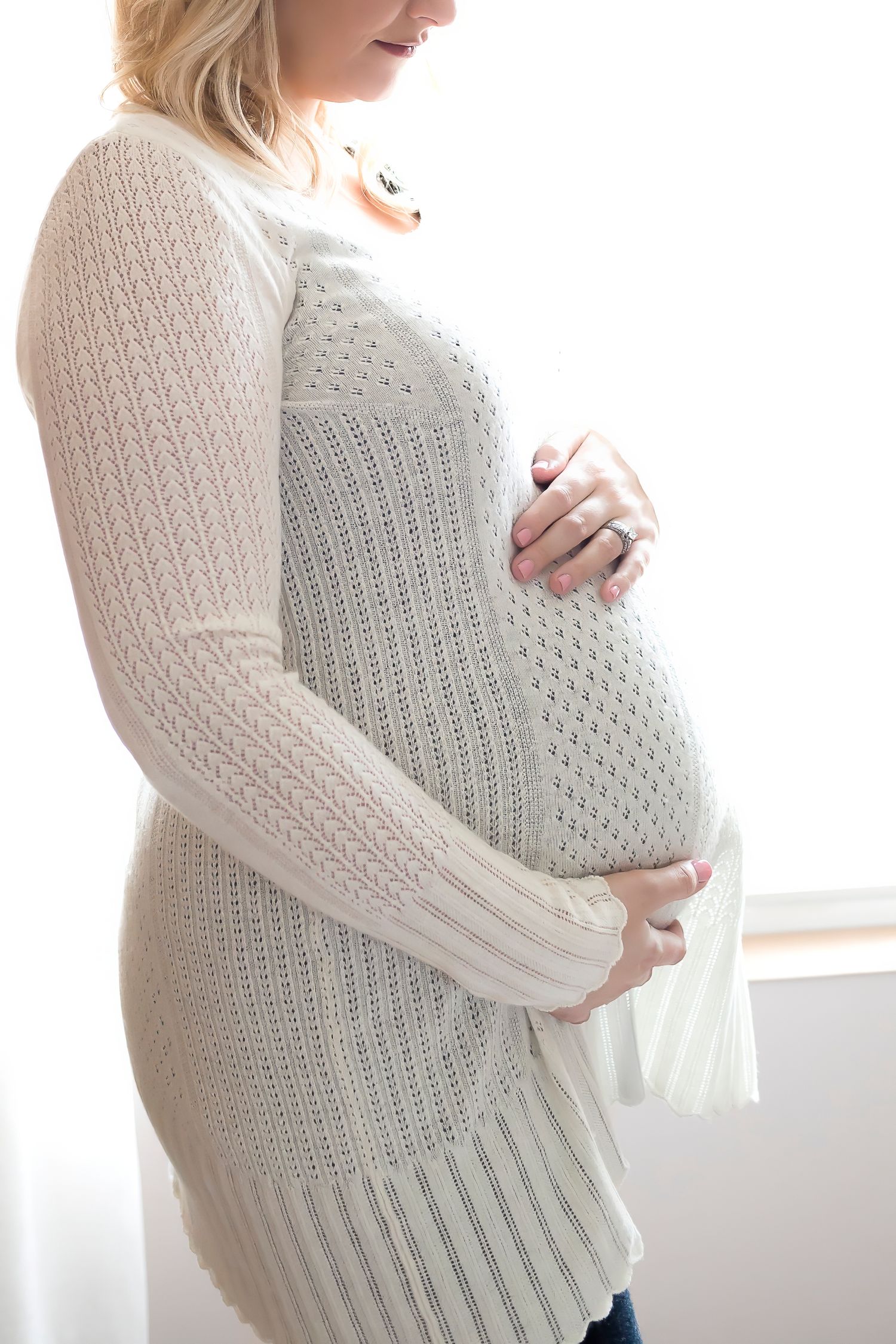 Expectant mother in a cozy white knit sweater gently cradles her baby bump by a window.