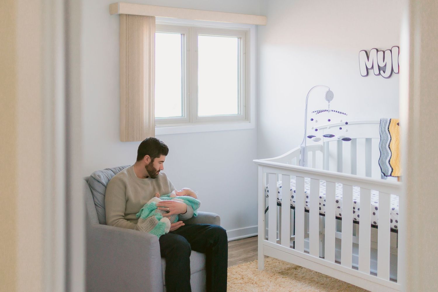 Father sitting in nursery chair holding newborn baby next to white crib in softly lit baby room.