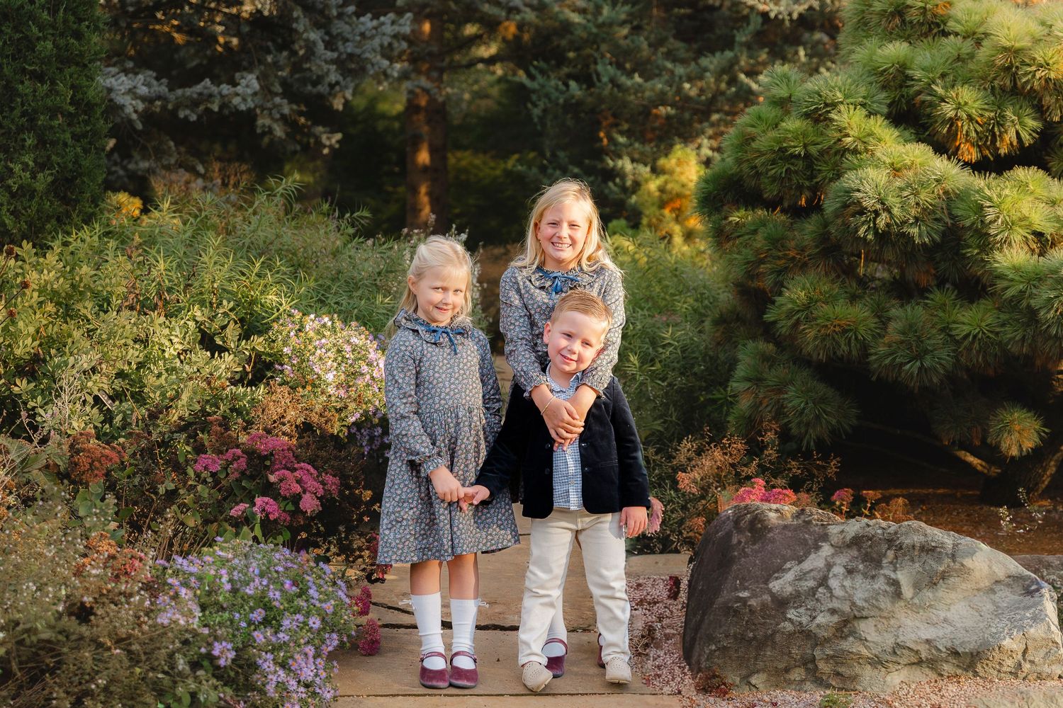 Three children smiling and posing together in a lush garden with colorful flowers and evergreen trees.