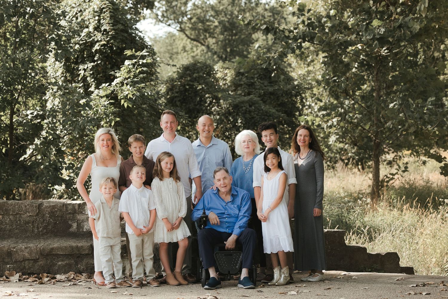 Large multigenerational family portrait outdoors, smiling adults and children posed together on stone steps surrounded by lush green trees.