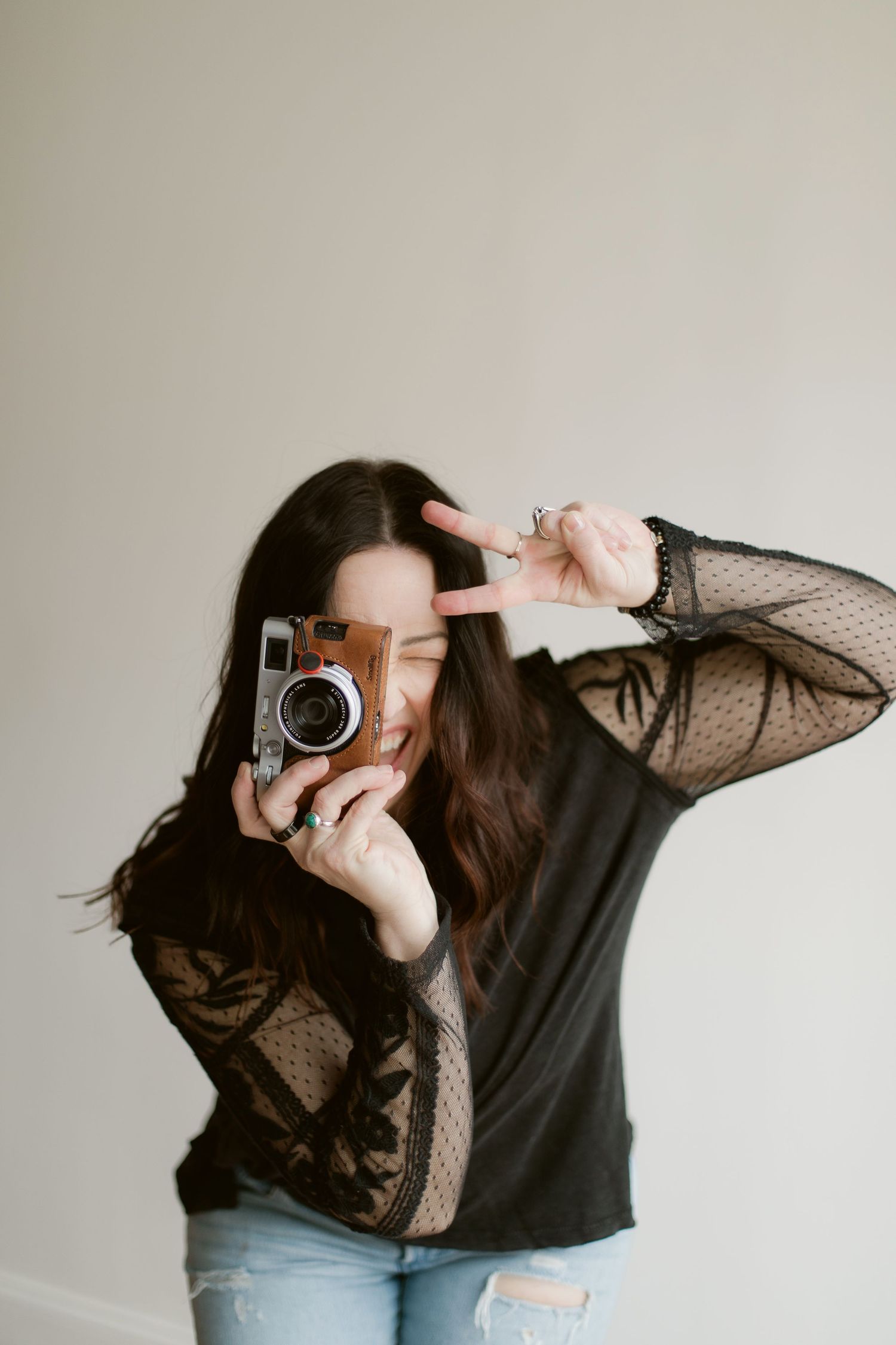 A photographer in a black lace sleeve top and jeans holds a vintage camera while making a peace sign gesture.
