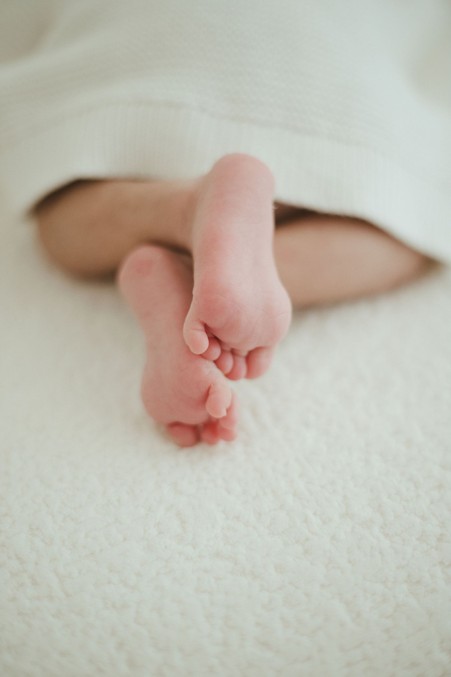 Tiny newborn baby feet peeking out from under a soft white blanket on a white textured surface.