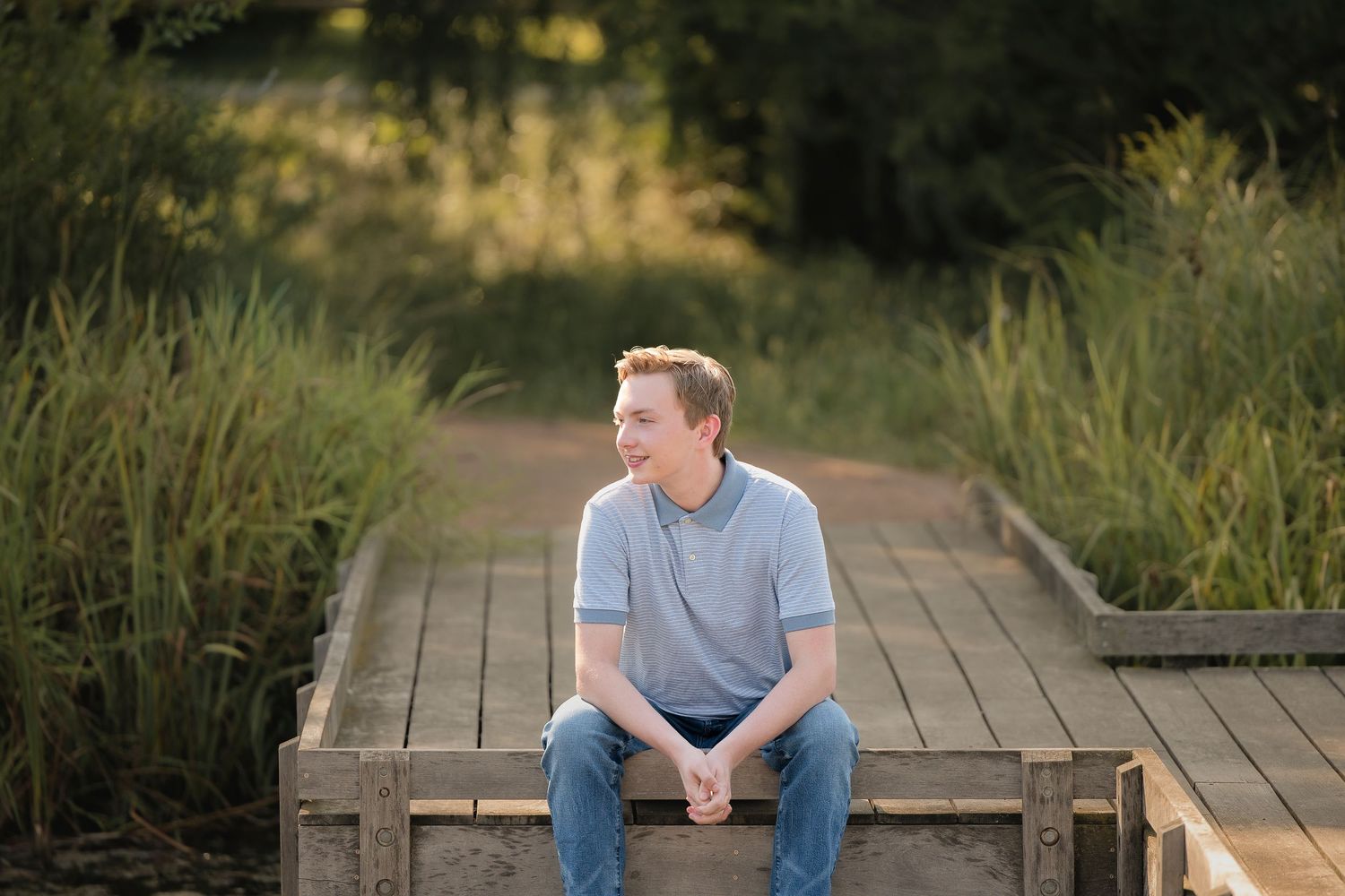 Young man with red hair sits on a wooden boardwalk surrounded by lush greenery in warm golden hour light.