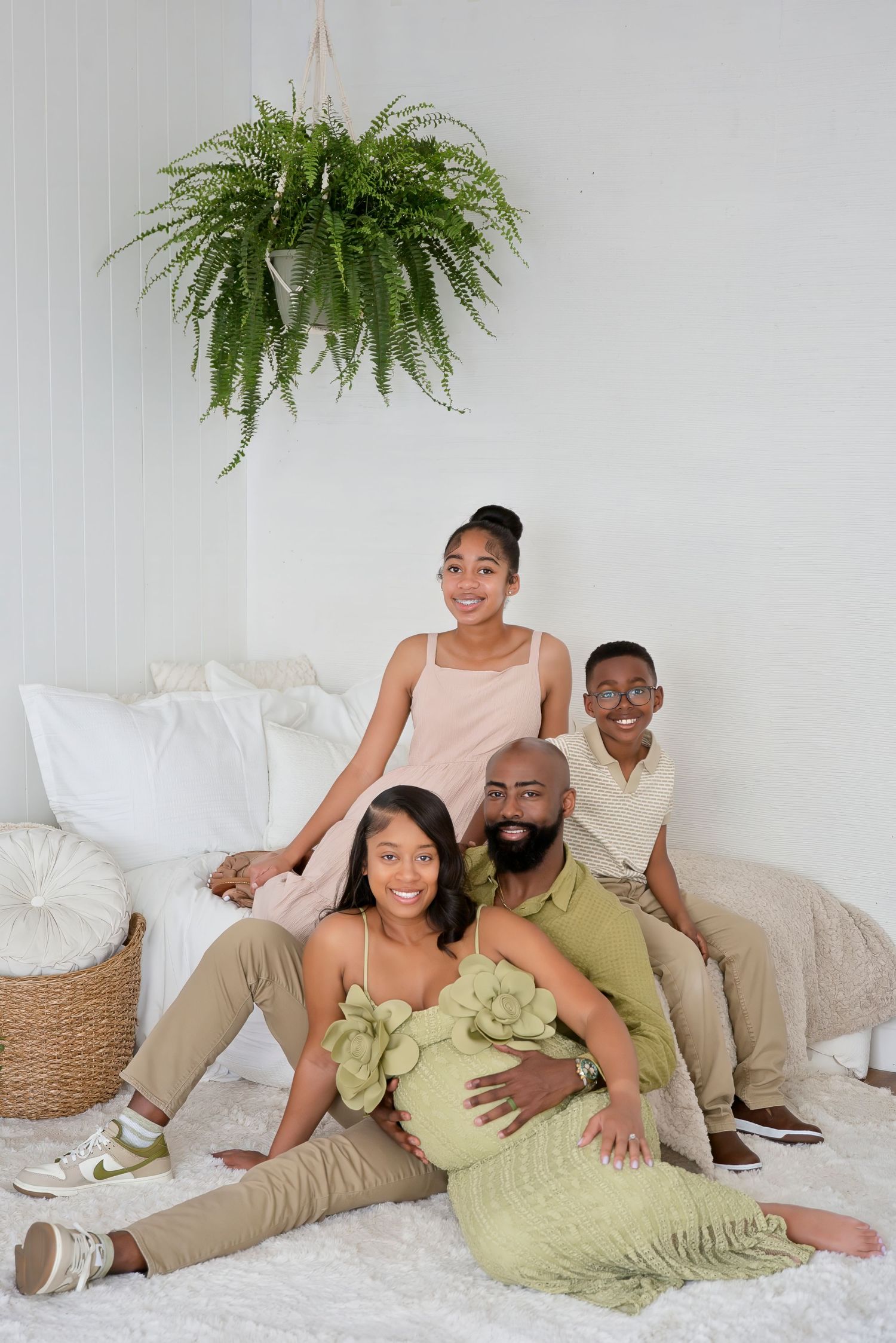 A happy Black family sits together on a white floor, with a pregnant woman in green, her bearded husband, and two teenagers.