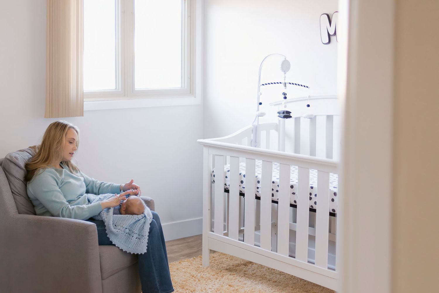Mother sitting in nursery chair holding newborn baby beside white crib with mobile in bright room.