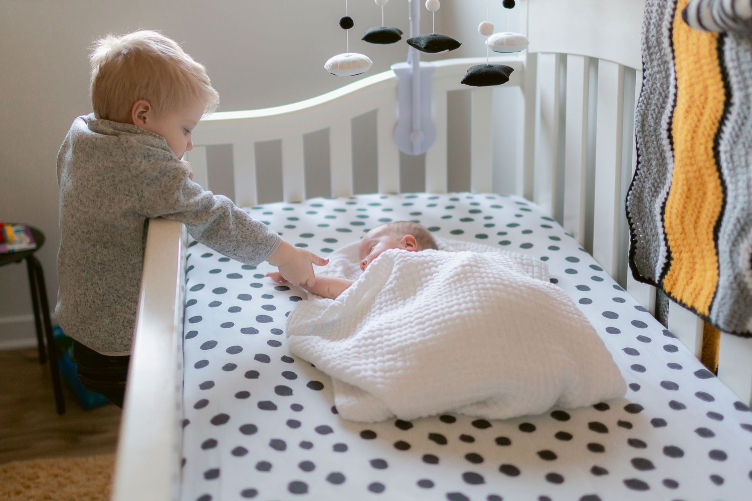 Toddler in gray hoodie leaning over white crib looking at sleeping newborn wrapped in white blanket.