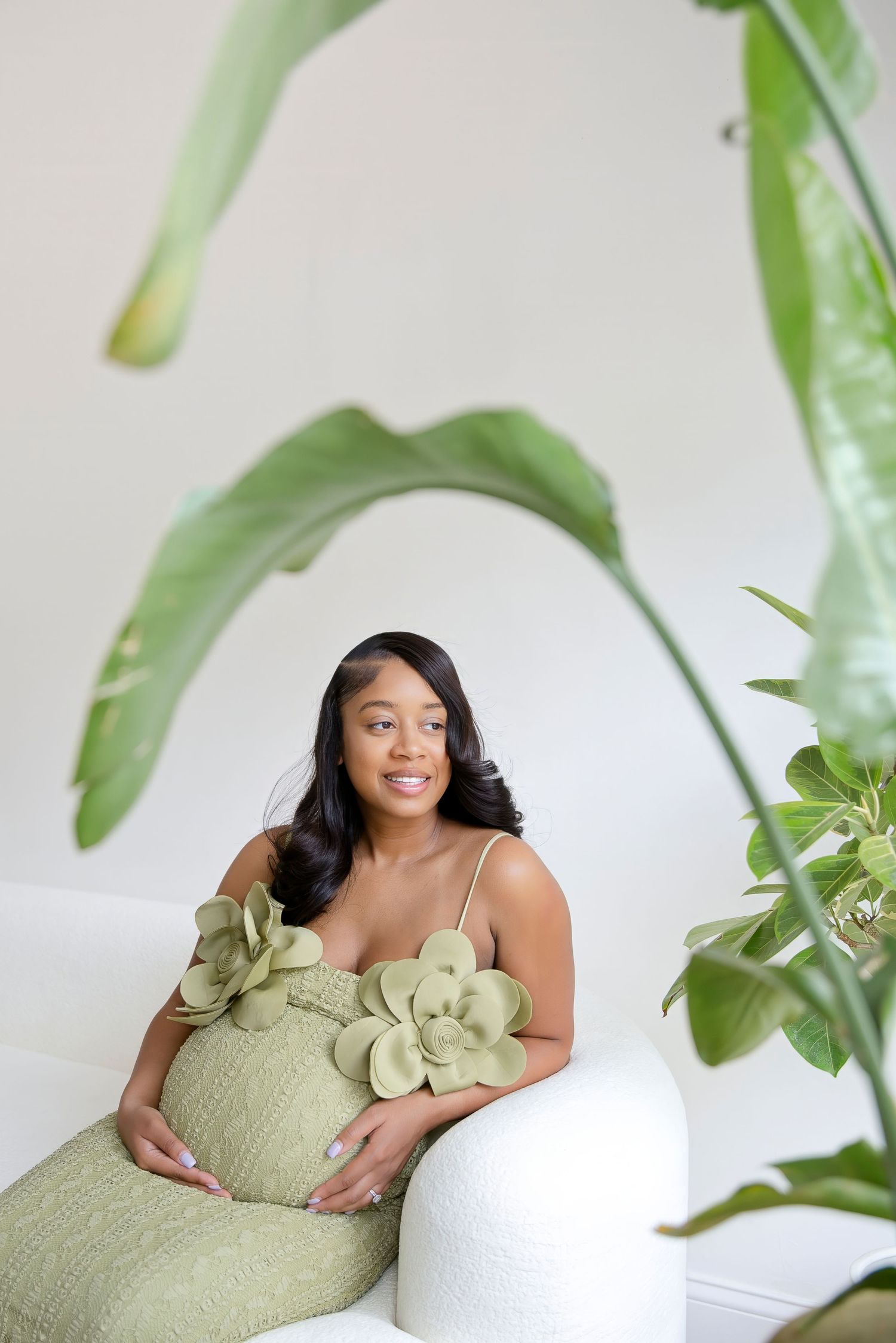 Pregnant woman in sage green floral dress sitting on white sofa surrounded by tropical plants.