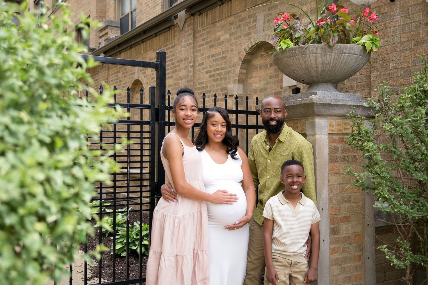 A joyful Black family poses outdoors, featuring a pregnant woman in white, a teen girl, a man, and a young boy.
