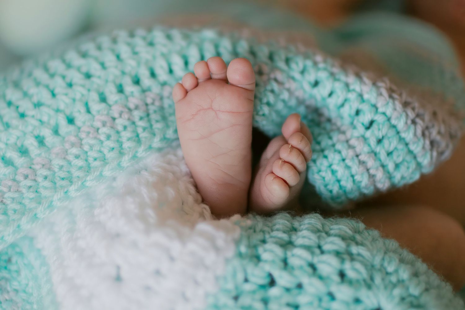 Tiny newborn baby feet peeking out from a soft mint green and white crocheted knit blanket.
