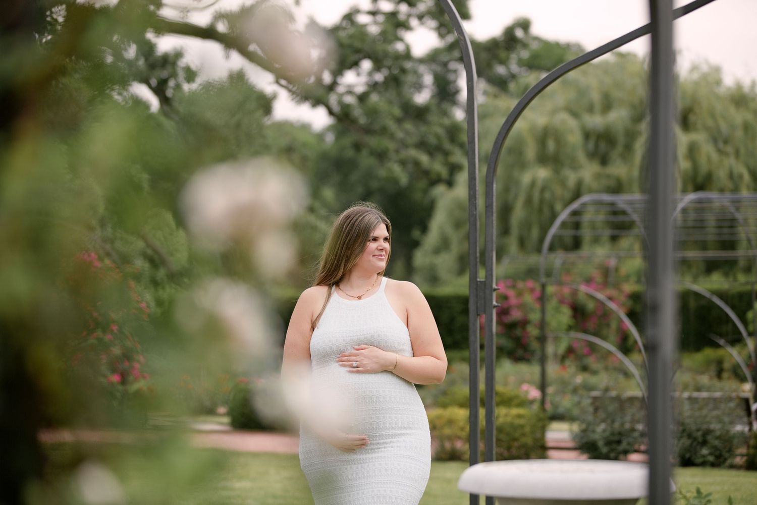 Pregnant woman in white dress cradling baby bump in lush garden with metal arch and blooming flowers.