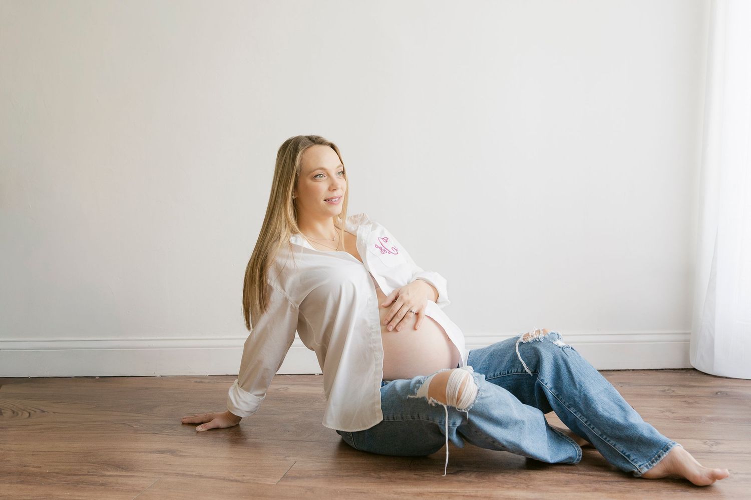 Pregnant woman in white blouse and ripped jeans sitting on hardwood floor cradling baby bump.