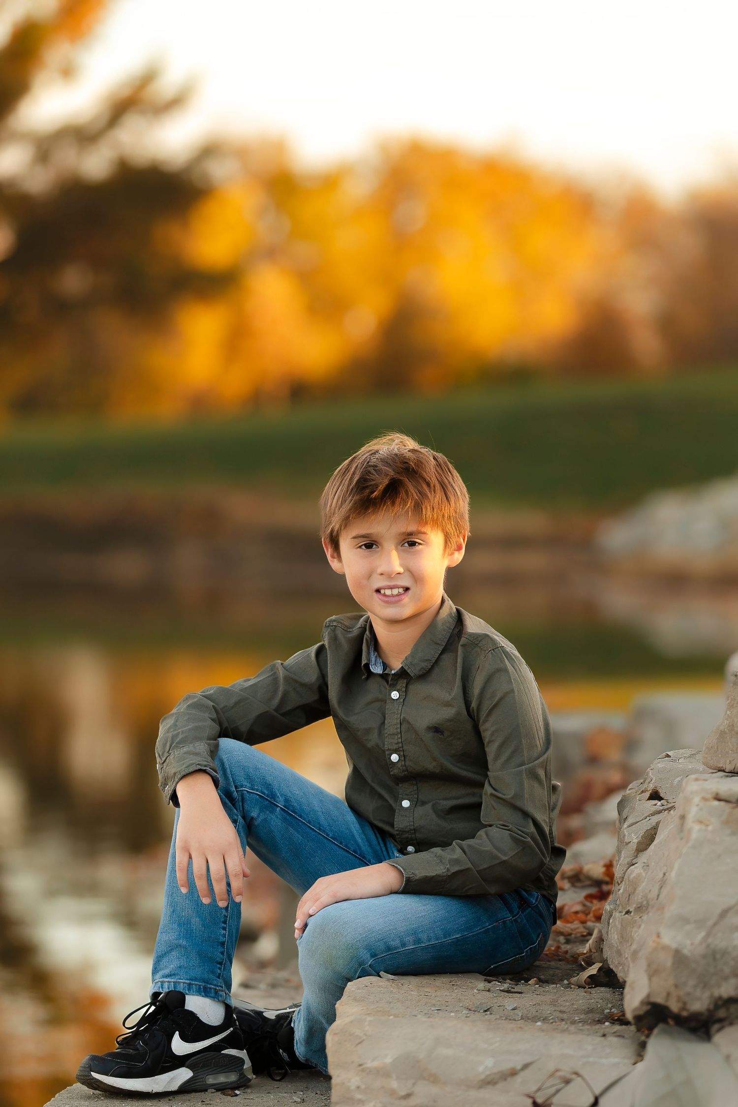 Person in green button-down shirt sits casually on rocky ledge with autumn foliage in background.