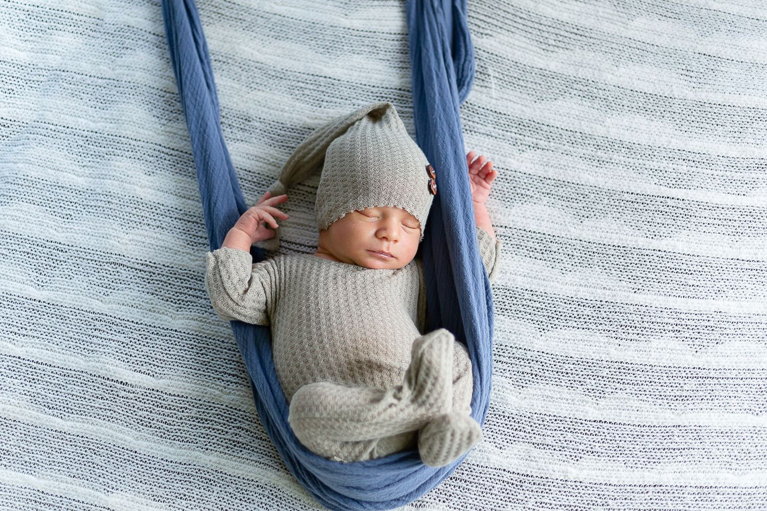 Newborn baby sleeping peacefully in a blue fabric hammock, wearing a gray knit outfit and matching hat.
