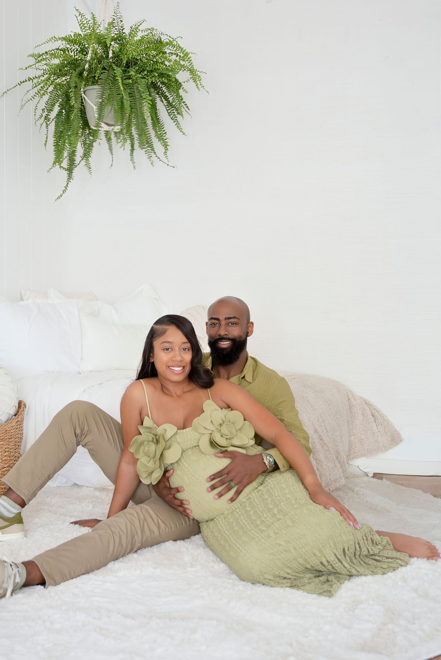 Pregnant woman in green floral dress sits with partner on white bed, both smiling warmly.