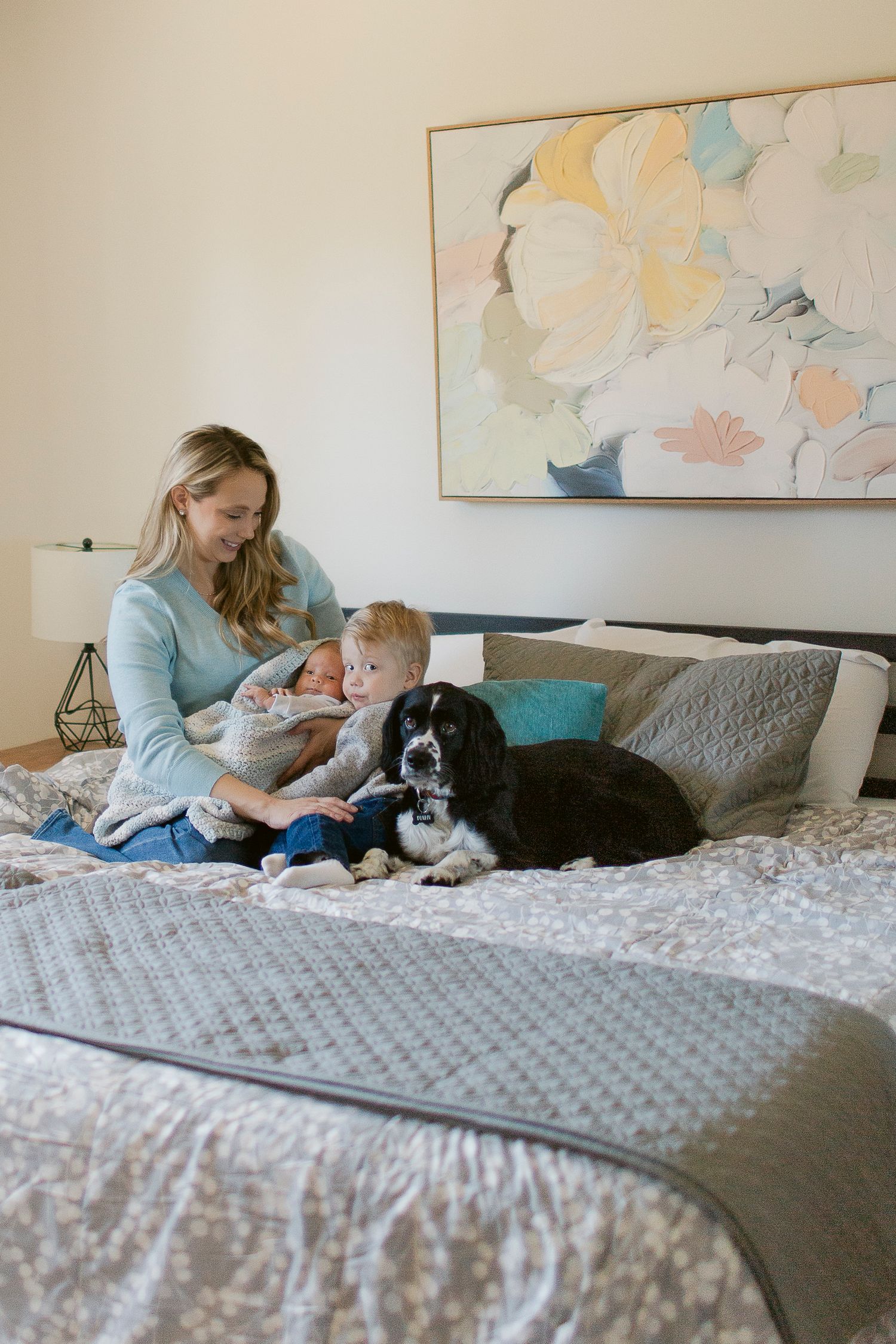 Mother with toddler and black spaniel dog relaxing on a cozy bed with floral artwork above.