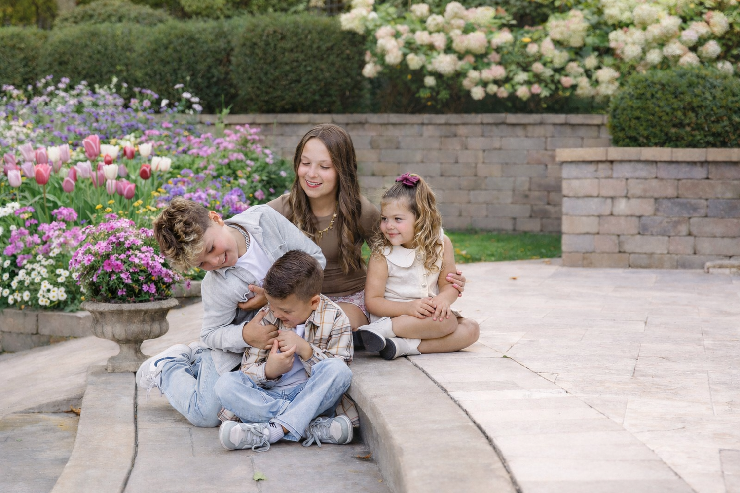 A group of people sit together on a stone walkway surrounded by blooming flowers in a garden setting.