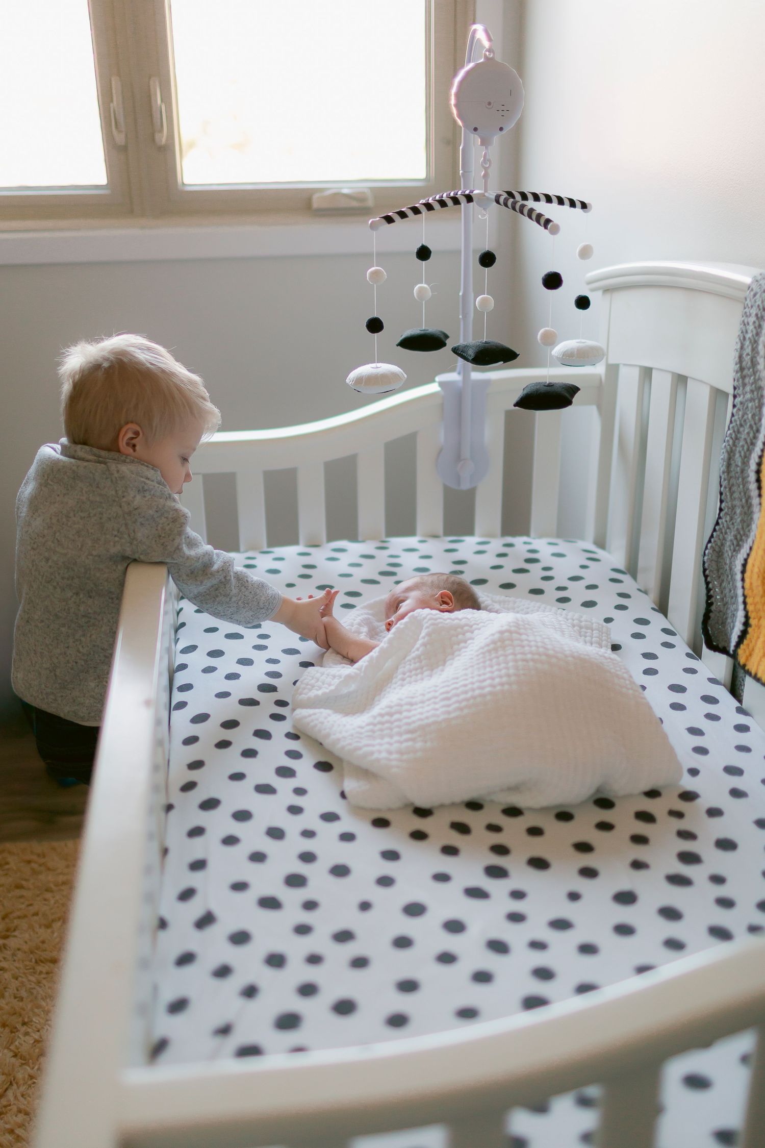 Toddler standing beside white crib with polka dot sheets, looking at sleeping newborn baby with mobile above.