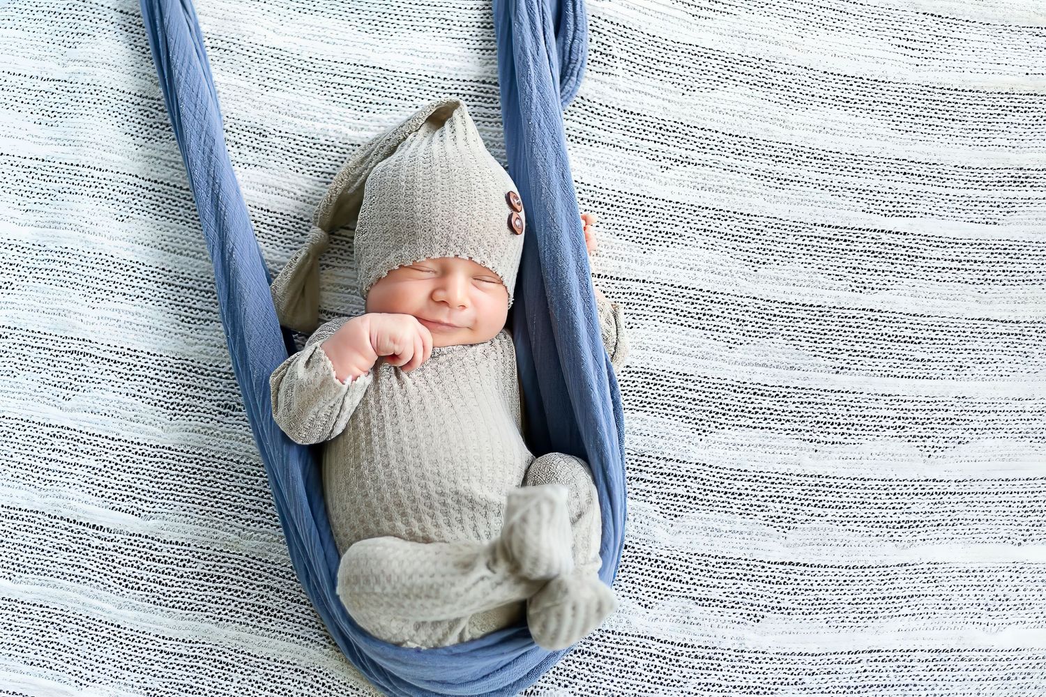 A tiny baby wearing a cream knit outfit rests peacefully in a blue fabric hammock against a striped background.
