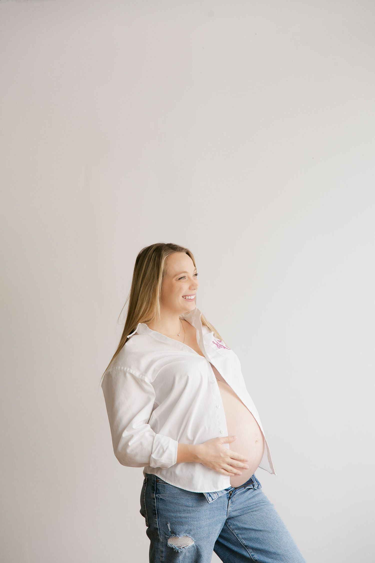 Smiling pregnant woman in white shirt and jeans cradling her baby bump against a light gray background.